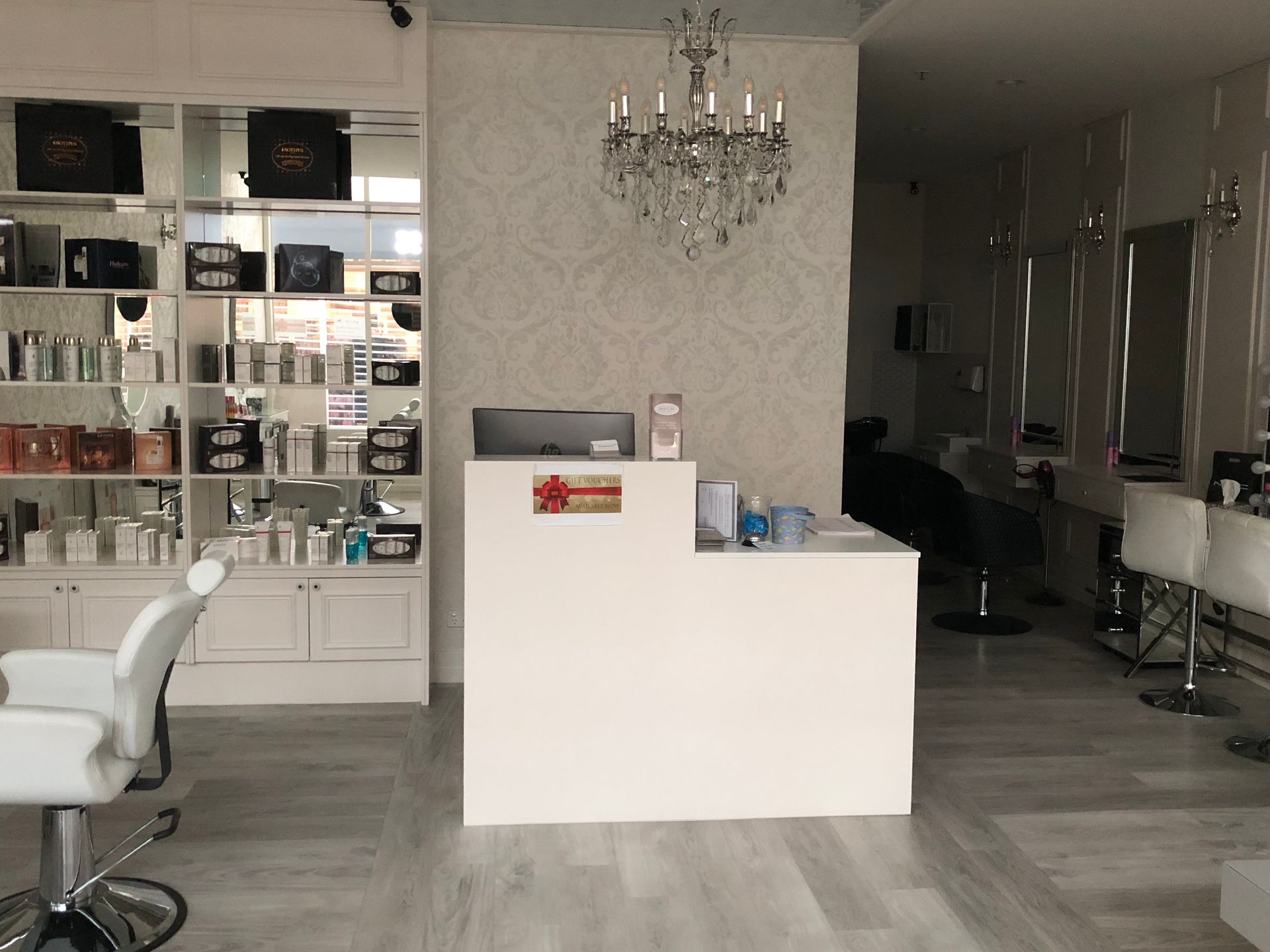Modern salon interior with white styling chairs, mirrored wall, reception desk, and chandelier.