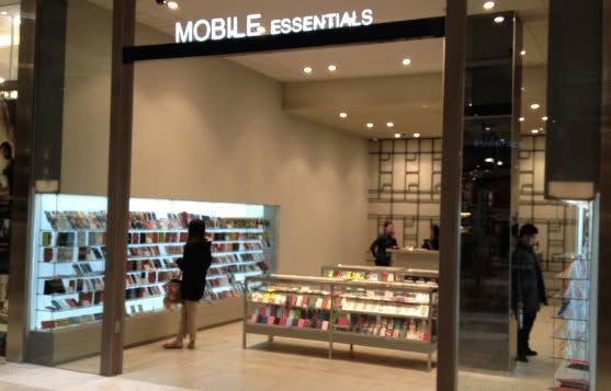 Storefront for “MOBILE ESSENTIALS” inside a mall, with illuminated product displays and a few shoppers.