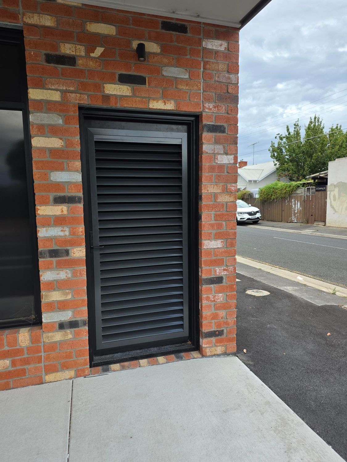 Black louvered door in a red brick building corner beside a sidewalk and street