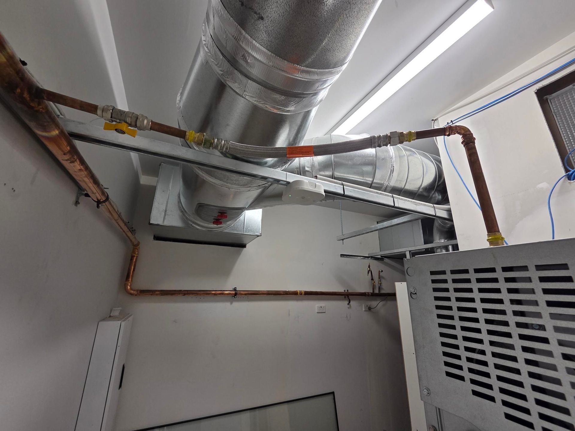 Exposed silver HVAC ducts and vents in a white utility room, with a large metal unit on the right.