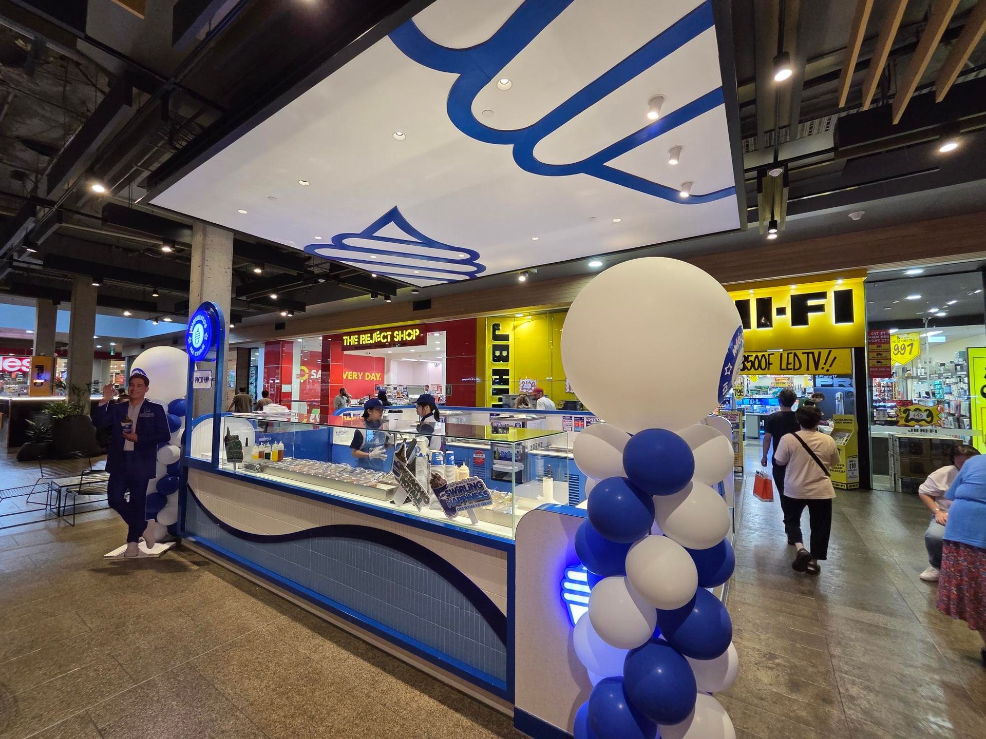 Kiosk with blue-and-white balloon arch in a shopping mall, with people browsing nearby