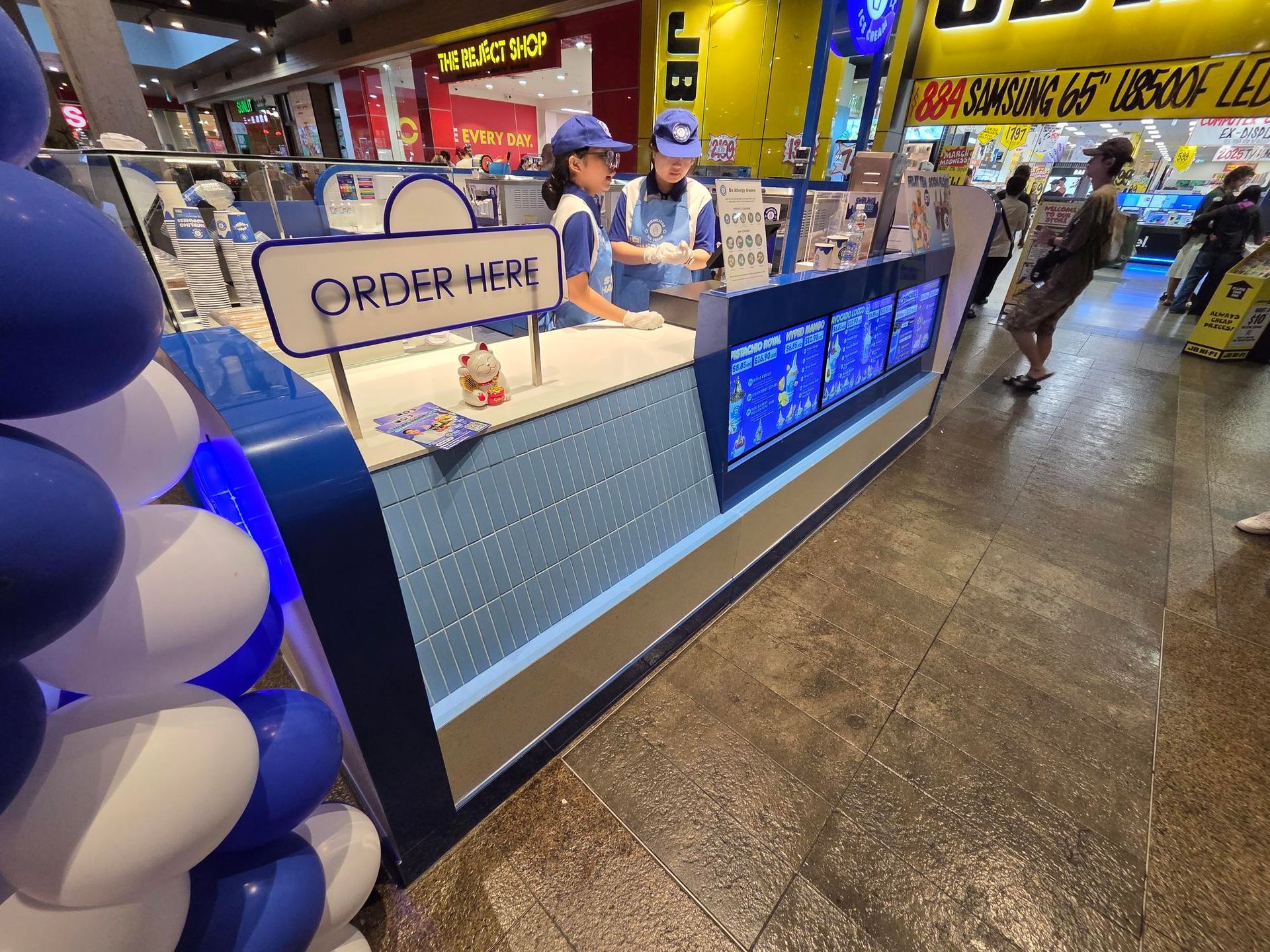 Coffee shop counter with “Order Here” sign, blue balloons, and staff serving customers in a mall.