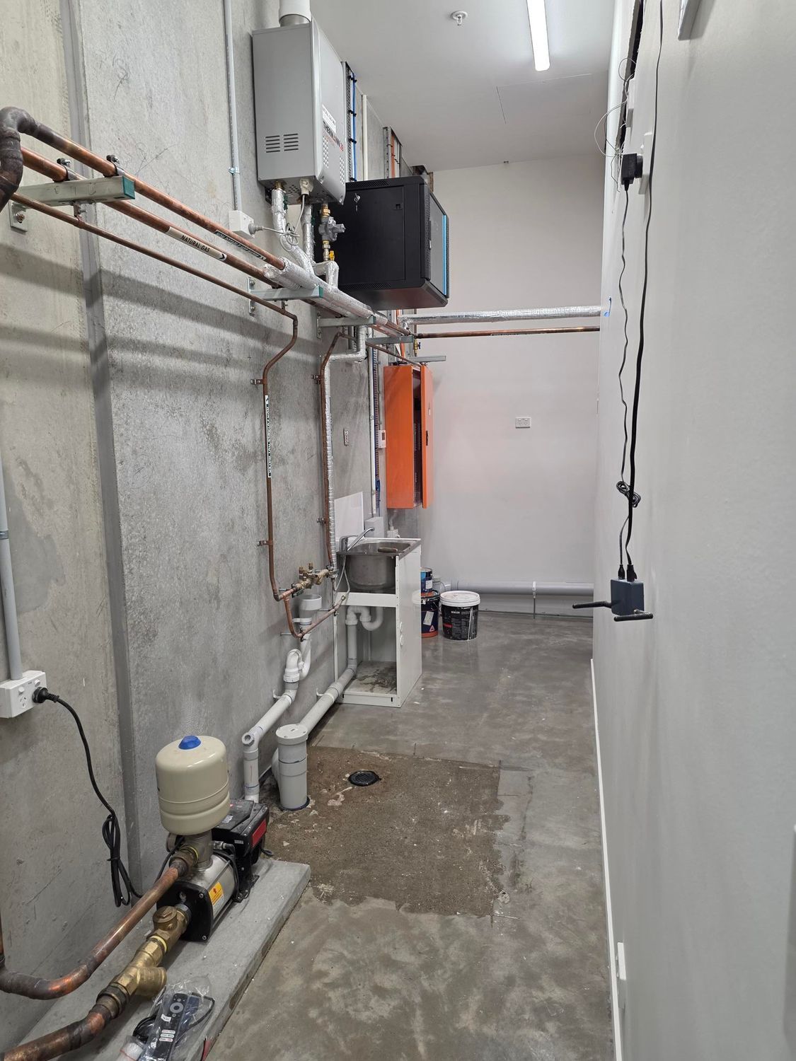 Narrow utility hallway with exposed pipes, valves, and equipment along gray concrete walls
