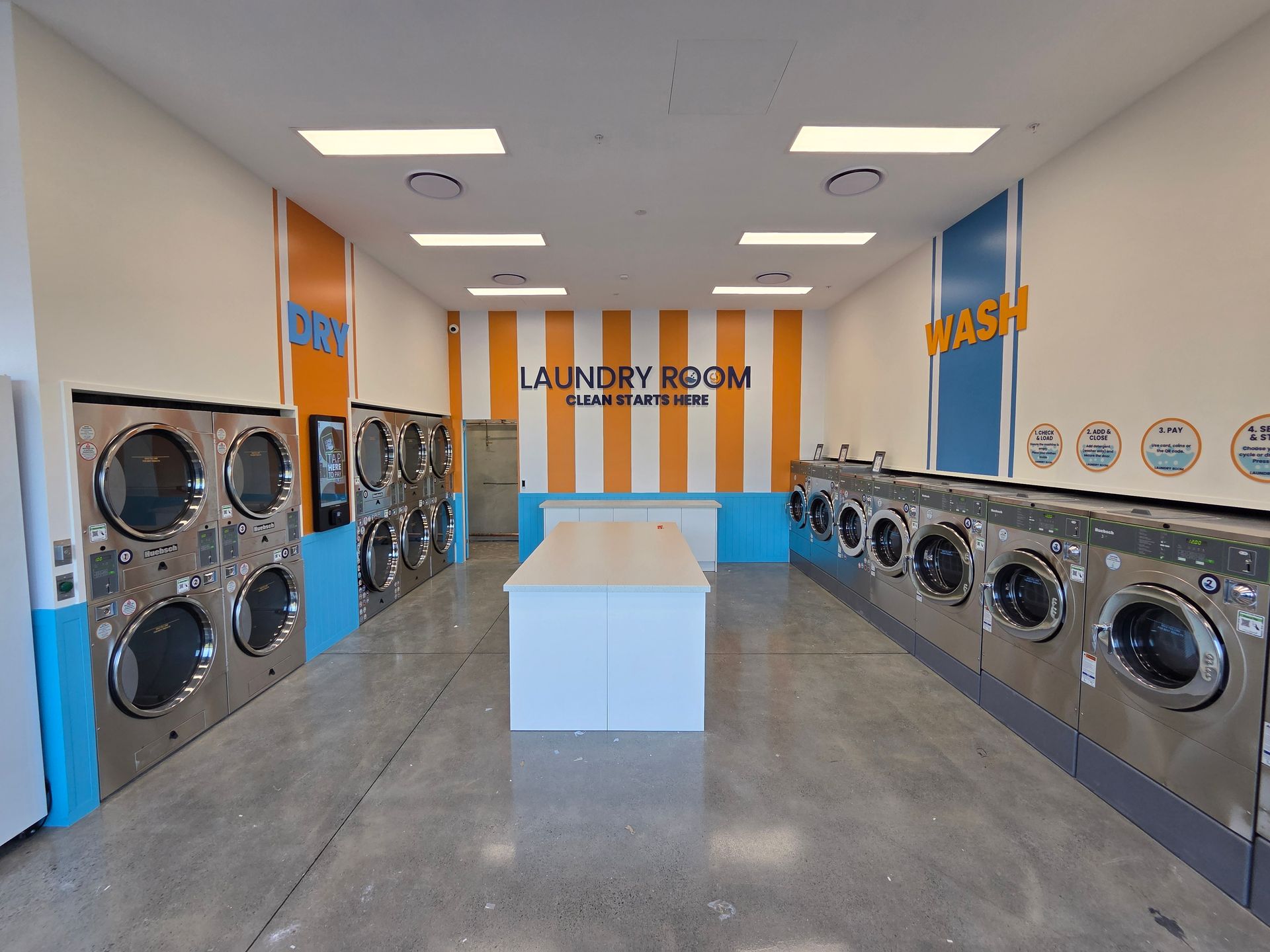 Bright laundromat with rows of washers and dryers, blue-and-orange walls, and a central folding table.