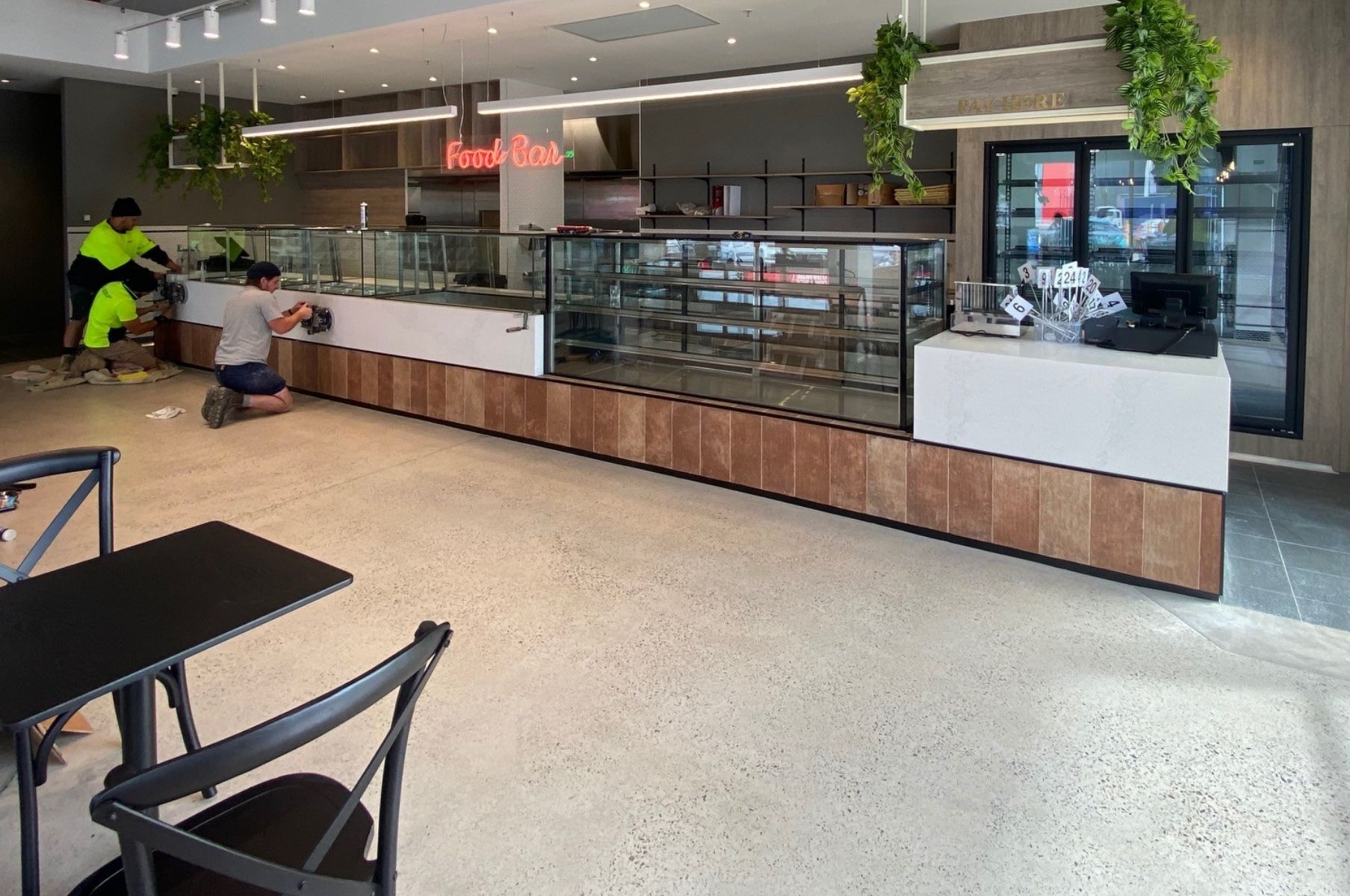Modern café counter with glass display case, hanging plants, and a few seated customers