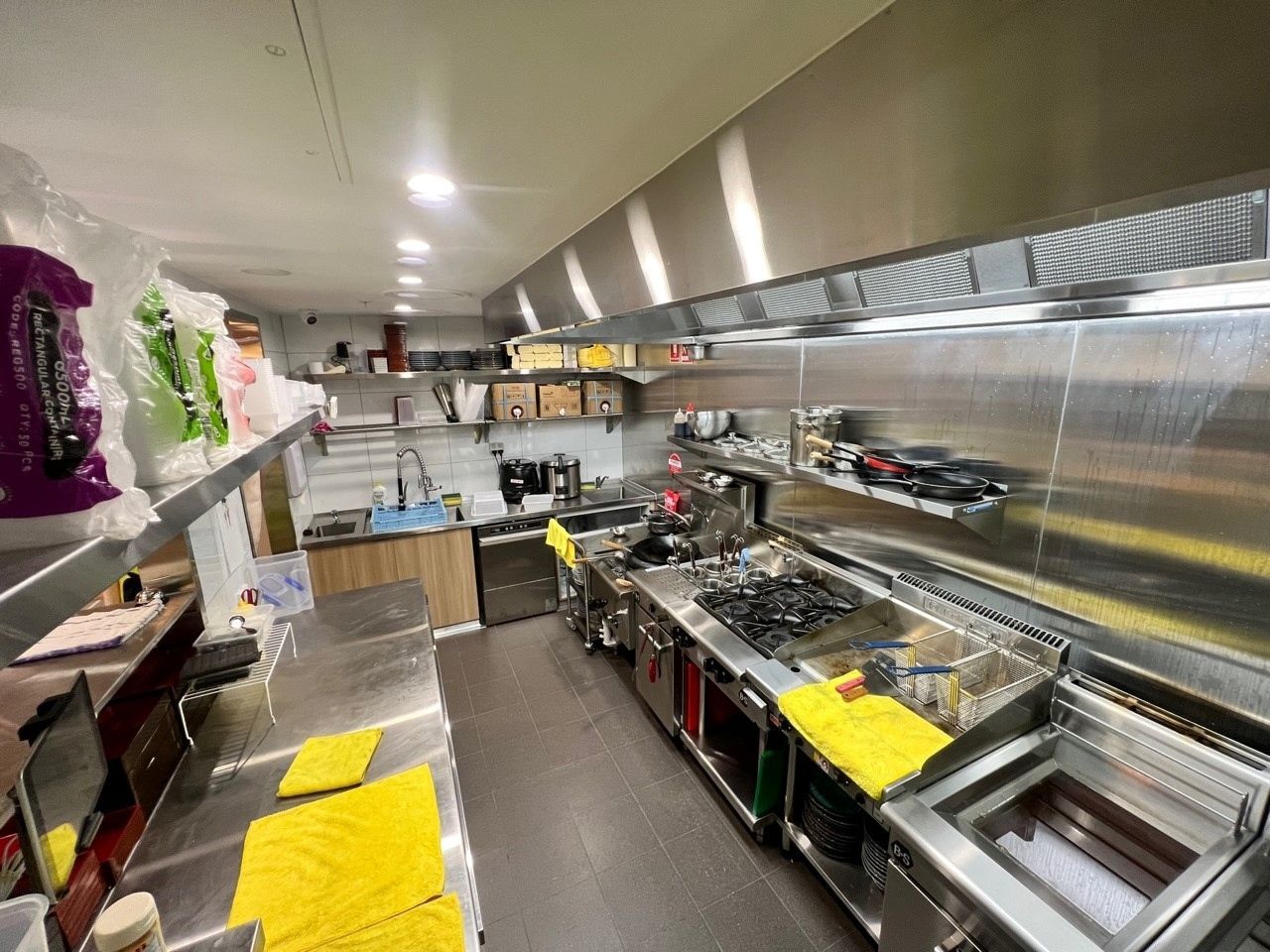 Commercial kitchen with stainless steel counters, cooking equipment, and yellow towels along a narrow prep area.