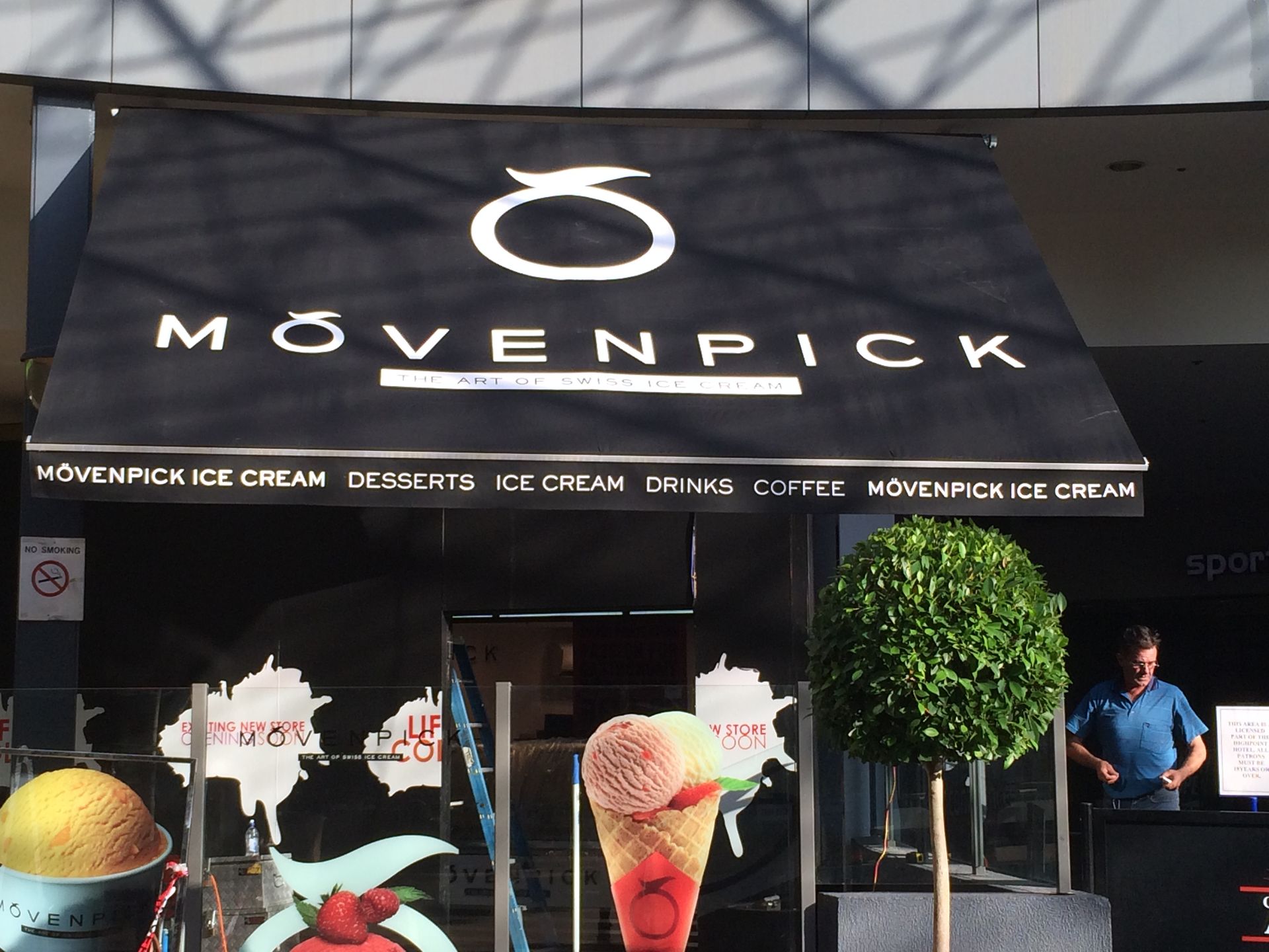 Mövenpick café storefront with black awning, ice cream cones, and two people standing outside
