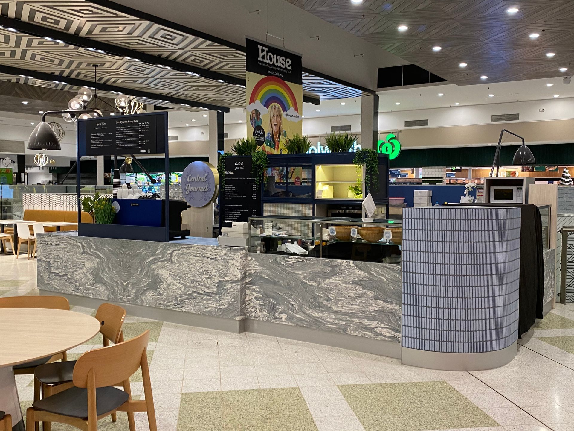 Indoor mall café kiosk with display cases, seating, and a rainbow sign behind the counter