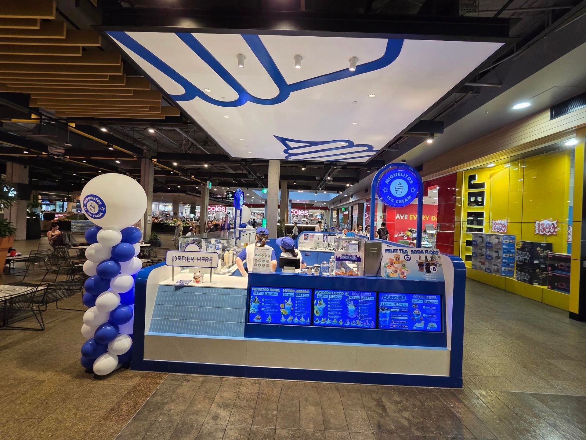 Blue-and-white kiosk counter in a shopping mall, with illuminated menu screens and balloon decorations