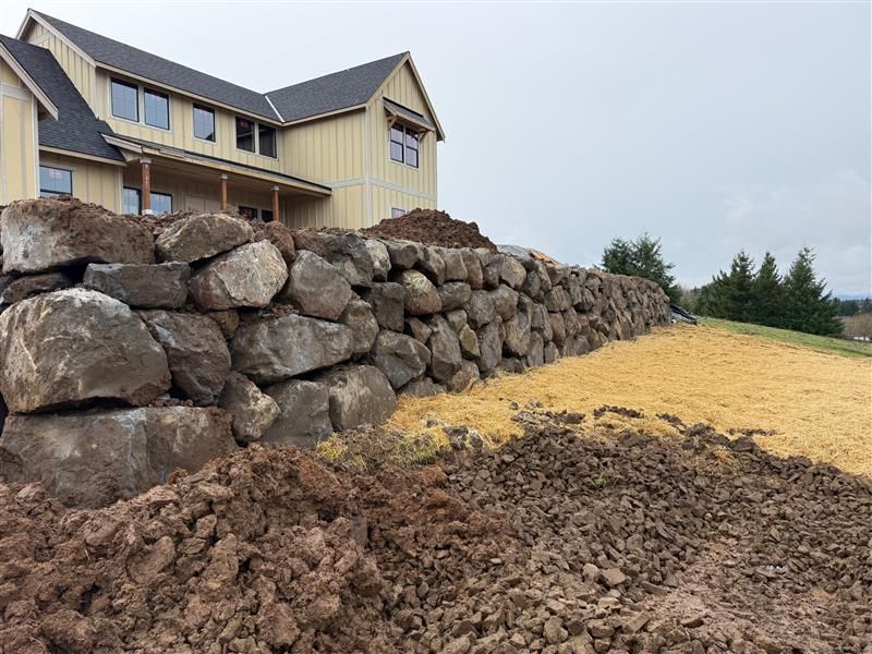 Stone retaining wall in front of a light yellow house with dark roof. Newly tilled earth.