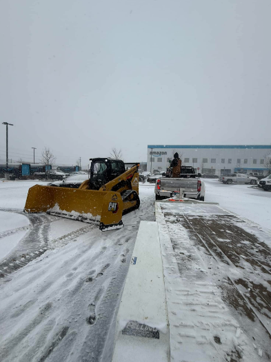 Snow plow clearing a snow-covered suburban street. Yellow blade pushes snow; white truck and houses in the background.