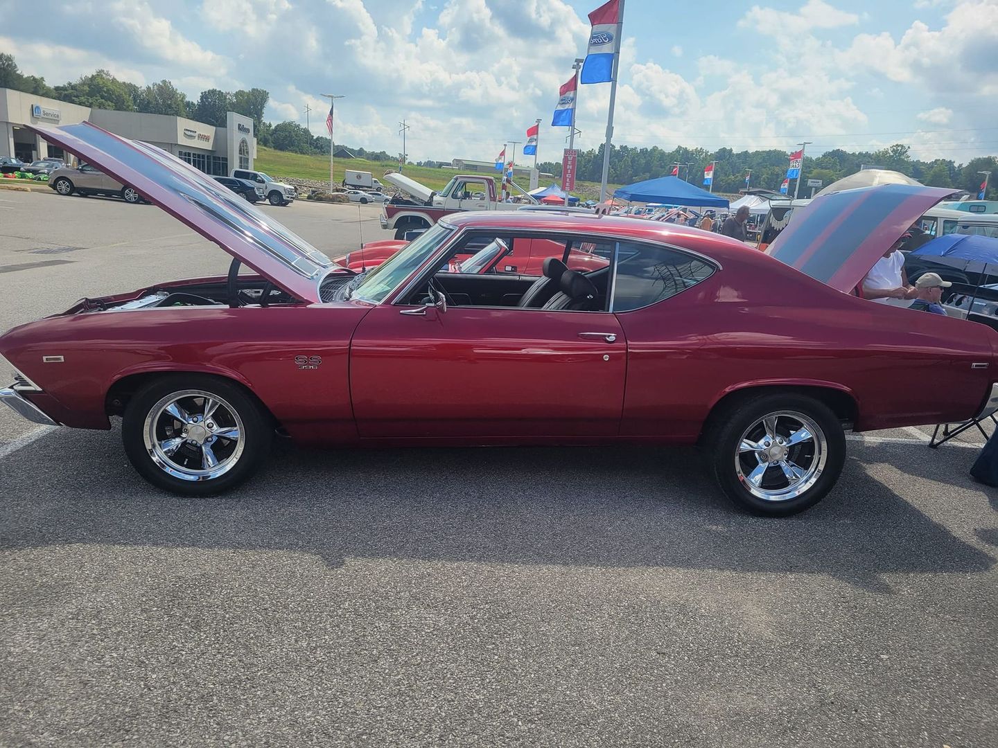 A cherry red Chevrolet Chevelle SS with its hood and trunk open, parked at an outdoor car show on a sunny day.