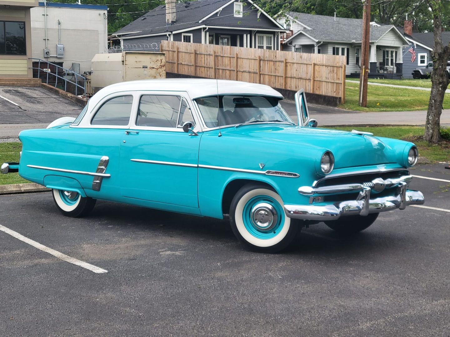 A turquoise and white vintage two-door sedan parked in a paved lot in front of houses.