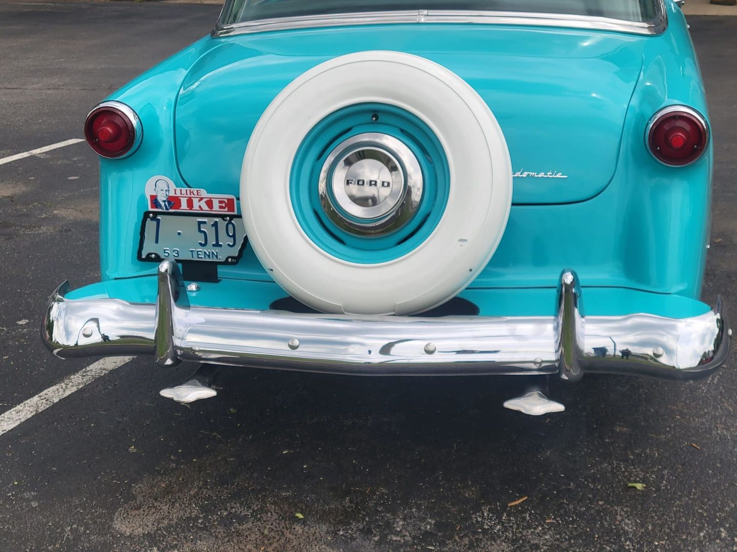 A turquoise vintage car rear view featuring a large, white-walled spare tire mounted on the trunk and a chrome bumper.