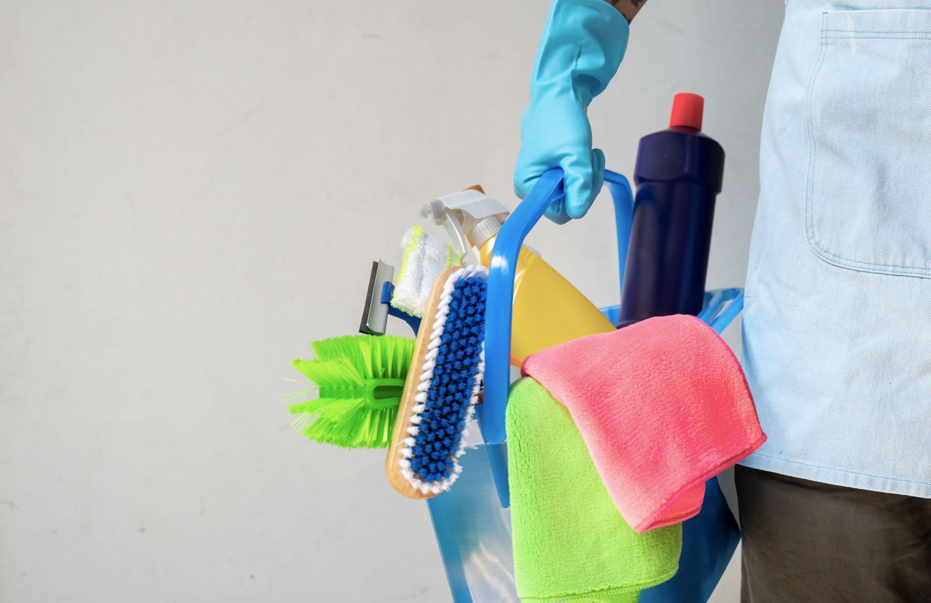 Person in blue gloves holds a blue cleaning bucket filled with supplies: brushes, cloths, and a bottle of cleaner.