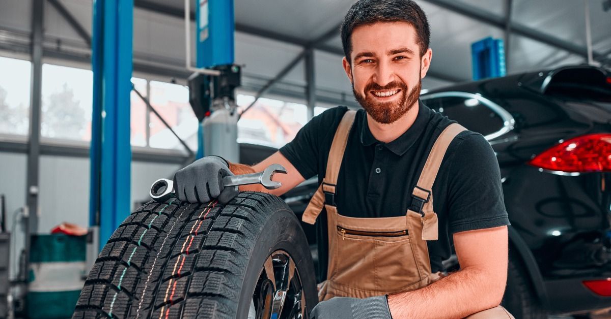 A technician holding a wrench on top of a tire beside a car in a garage.