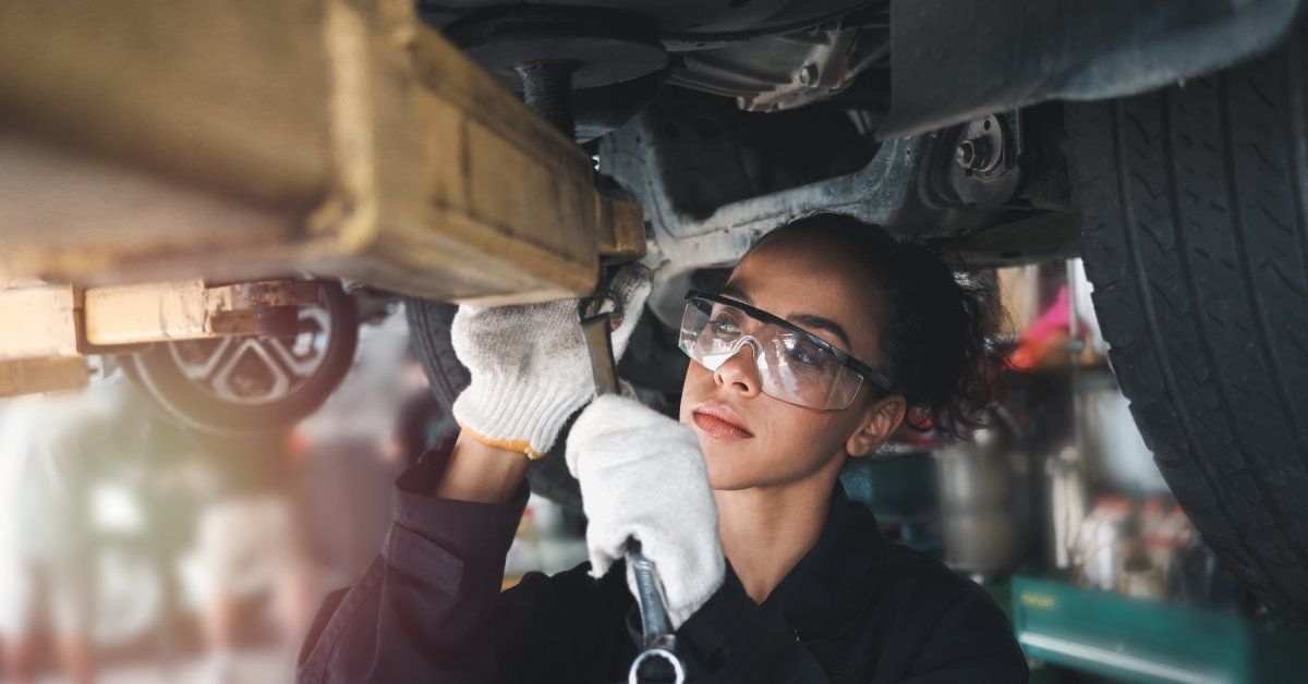 A woman wearing goggles uses a tool to adjust a part under a lifted vehicle.