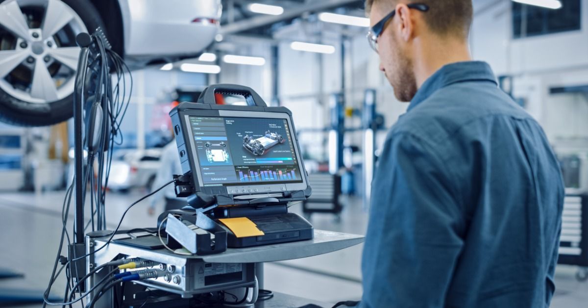 A technician using a diagnostic computer next to a raised car inside a shop.