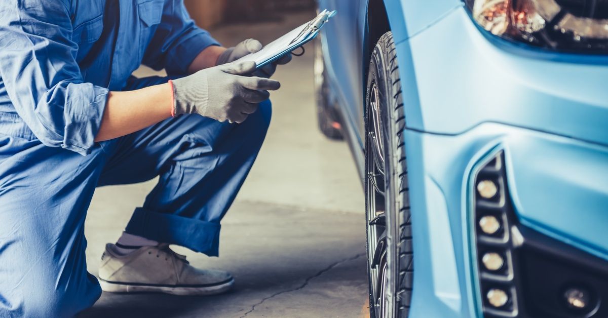 A mechanic is crouching and inspecting a car's tires while holding a clipboard.