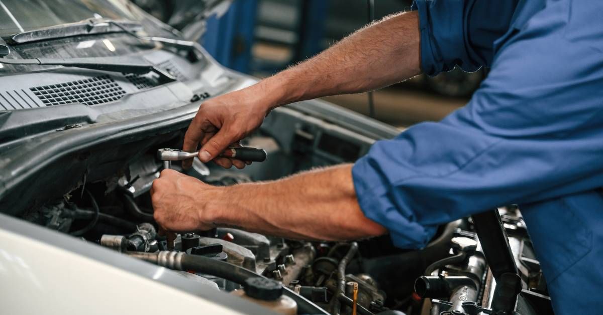 A man in a blue shirt uses a socket wrench on a car engine under an open hood inside a repair garage.