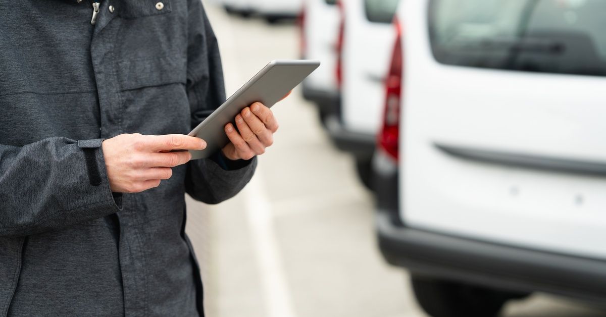 A person holding a tablet beside a row of parked white cargo vans outside.