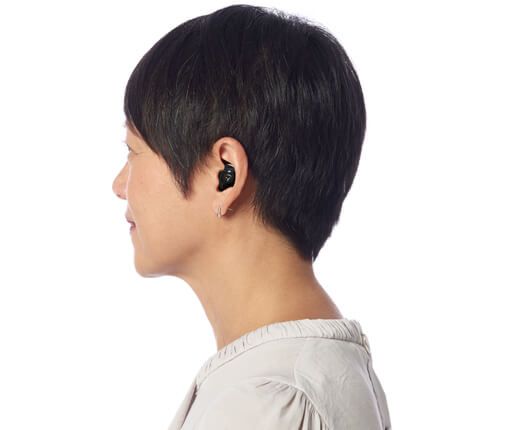 Woman with black in-ear hearing aid, side profile. Short, dark hair. Wearing a light-colored top.