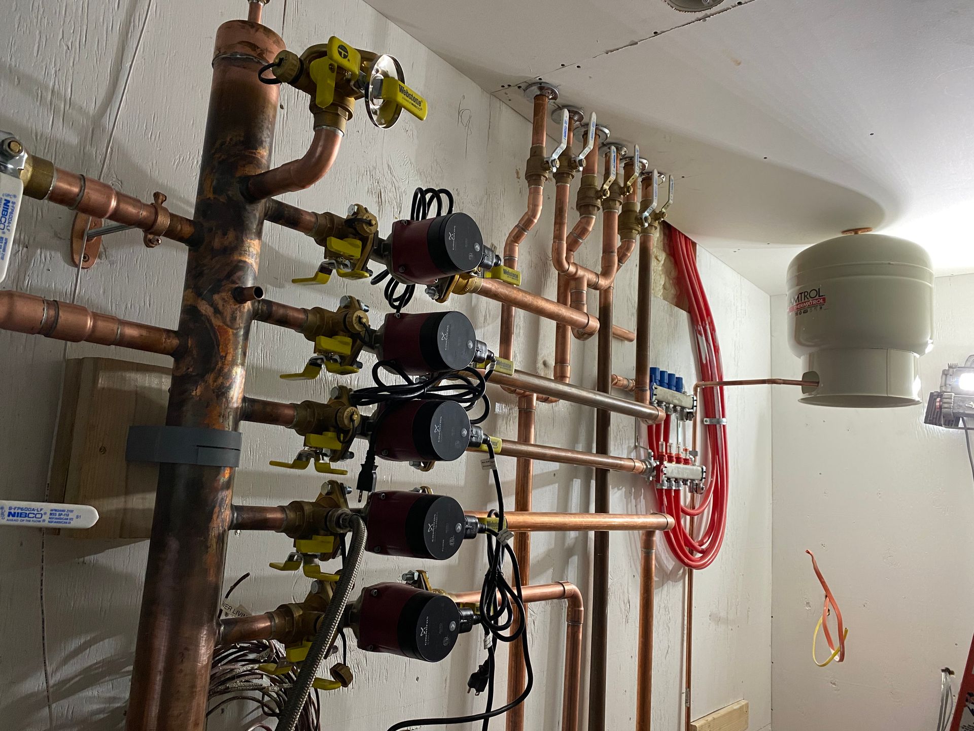 Copper pipes and valves in a utility room, with a red coil and white expansion tank on the wall.