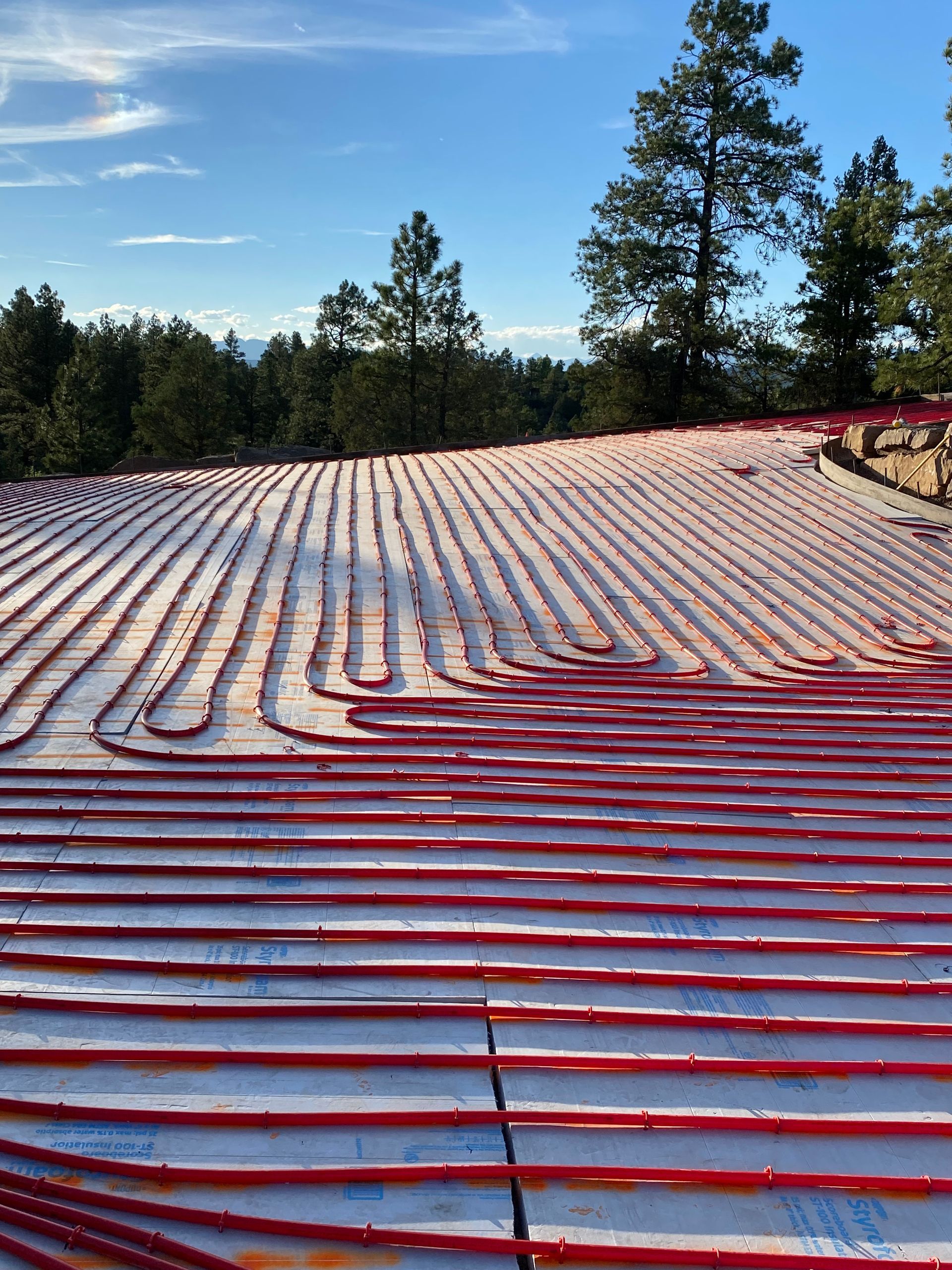 Snow-covered red roof with rows of white solar panels under a blue sky and pine trees