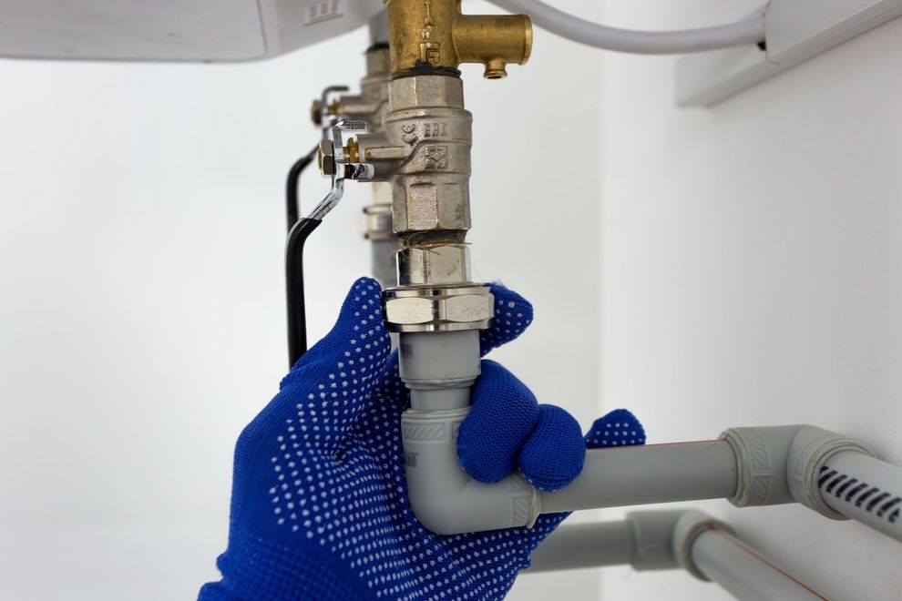 Blue-gloved hand adjusting a plumbing valve under a sink, with gray and brass pipes.