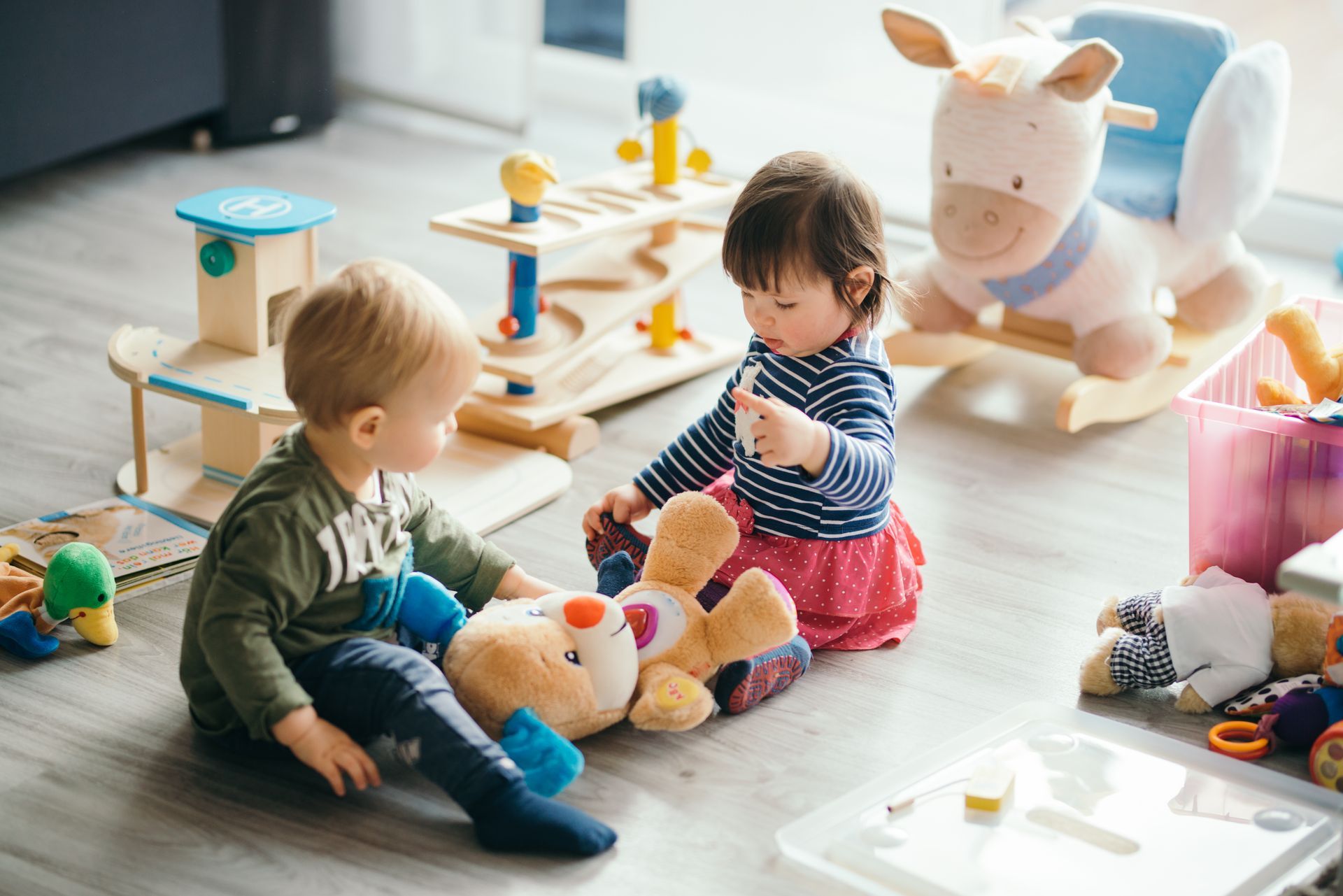 Two children playing with stuffed animals on a bright nursery floor near a toy cow and wooden toys