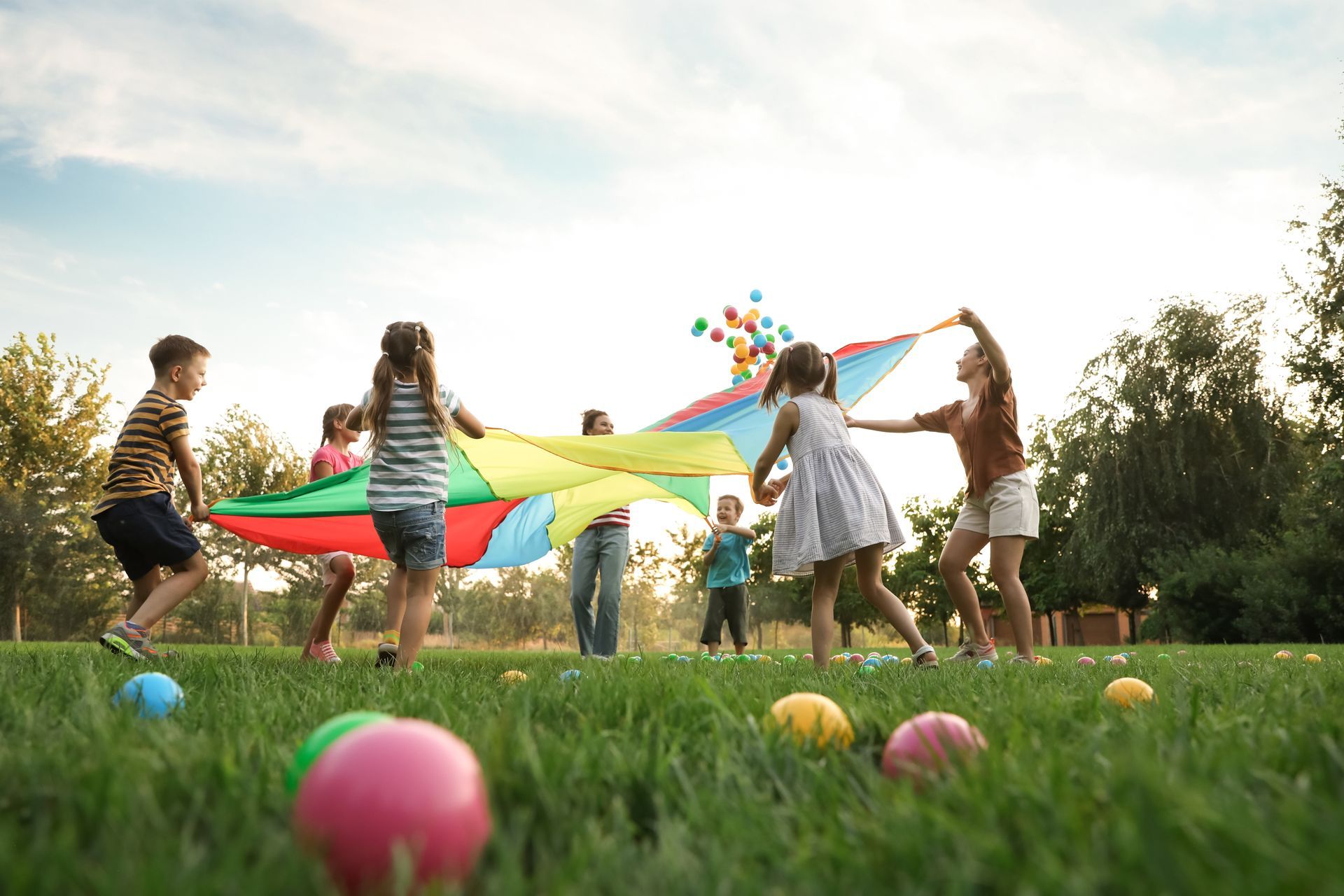 Children playing tug-of-war with a colorful parachute in a grassy park, with balloons scattered nearby.
