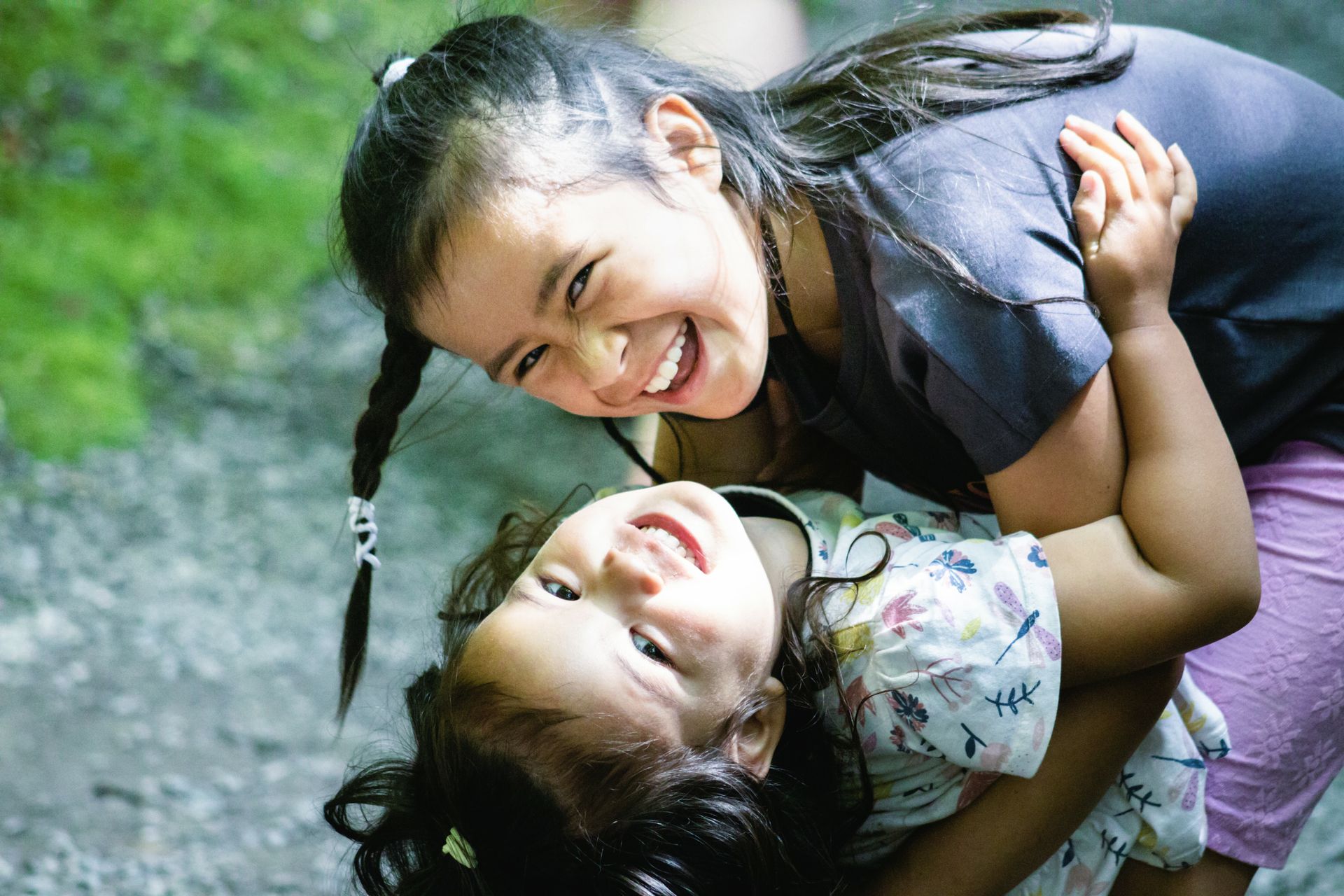Two smiling children lying on grass, playfully leaning over each other outdoors.