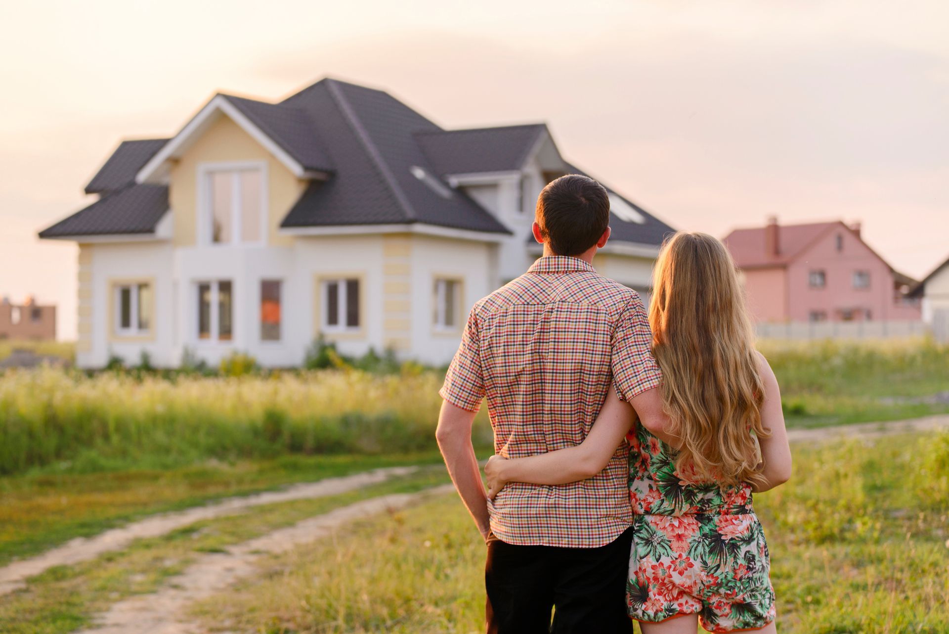 A couple with arms around each other looking at a suburban house during sunset in a grassy field. A couple with arms around each other looking at a suburban house during sunset in a grassy field.