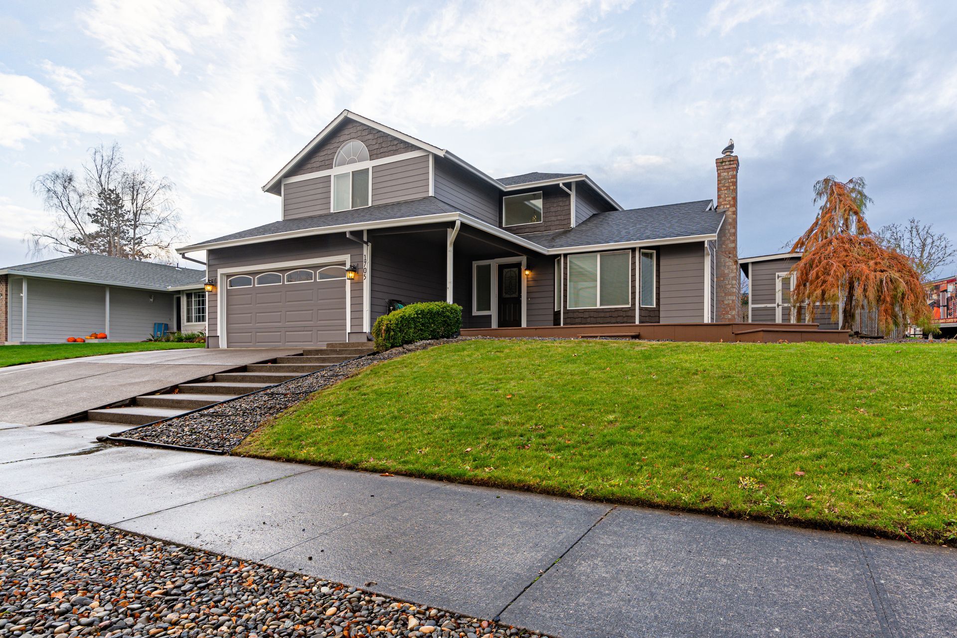 A two-story, gray-sided house with a stone chimney, a green lawn, and a wide driveway under a cloudy sky. A two-story, gray-sided house with a stone chimney, a green lawn, and a wide driveway under a cloudy sky.