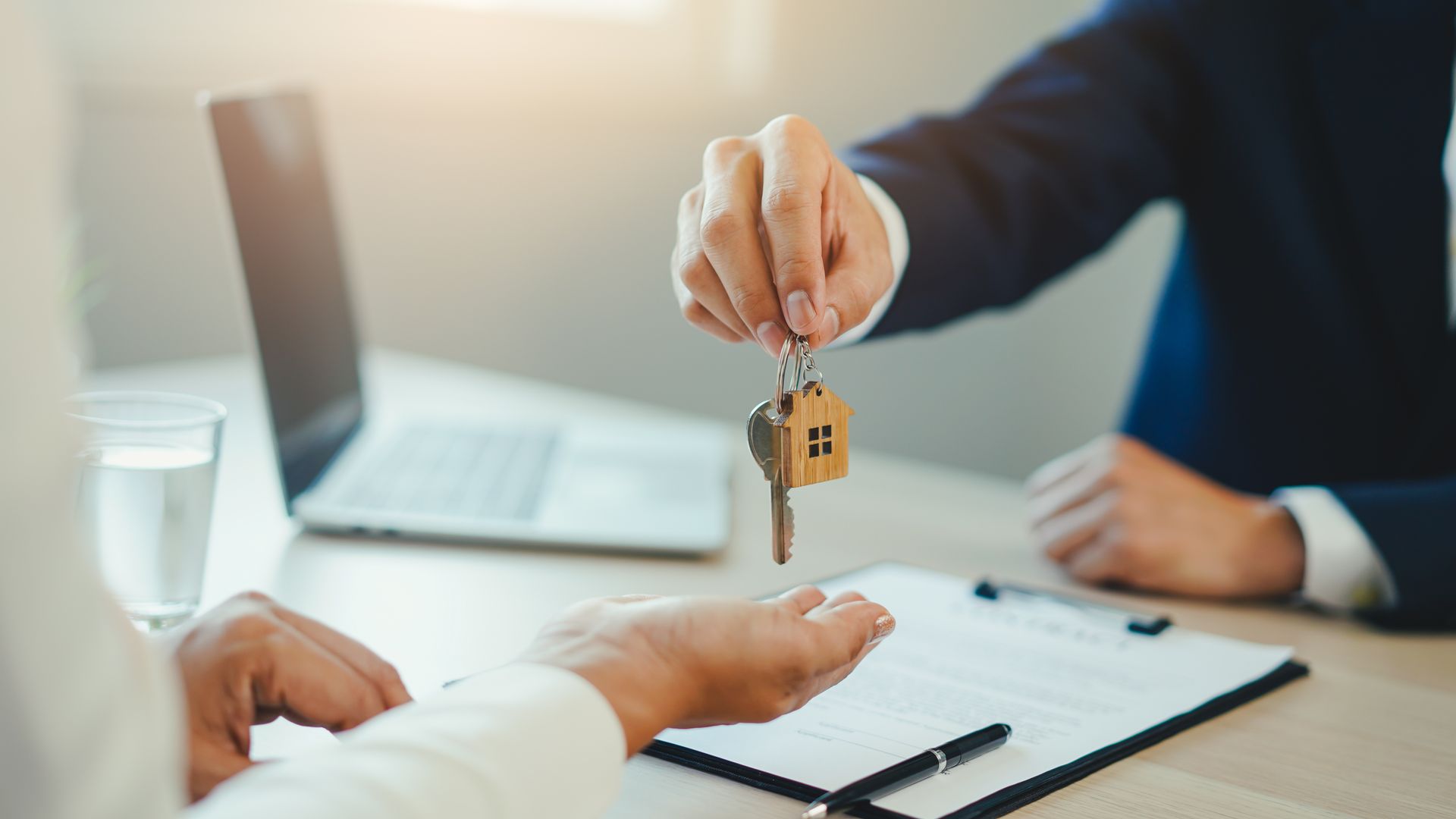 A person in a business suit hands keys with a house-shaped keychain to another person at an office desk. A person in a business suit hands keys with a house-shaped keychain to another person at an office desk.