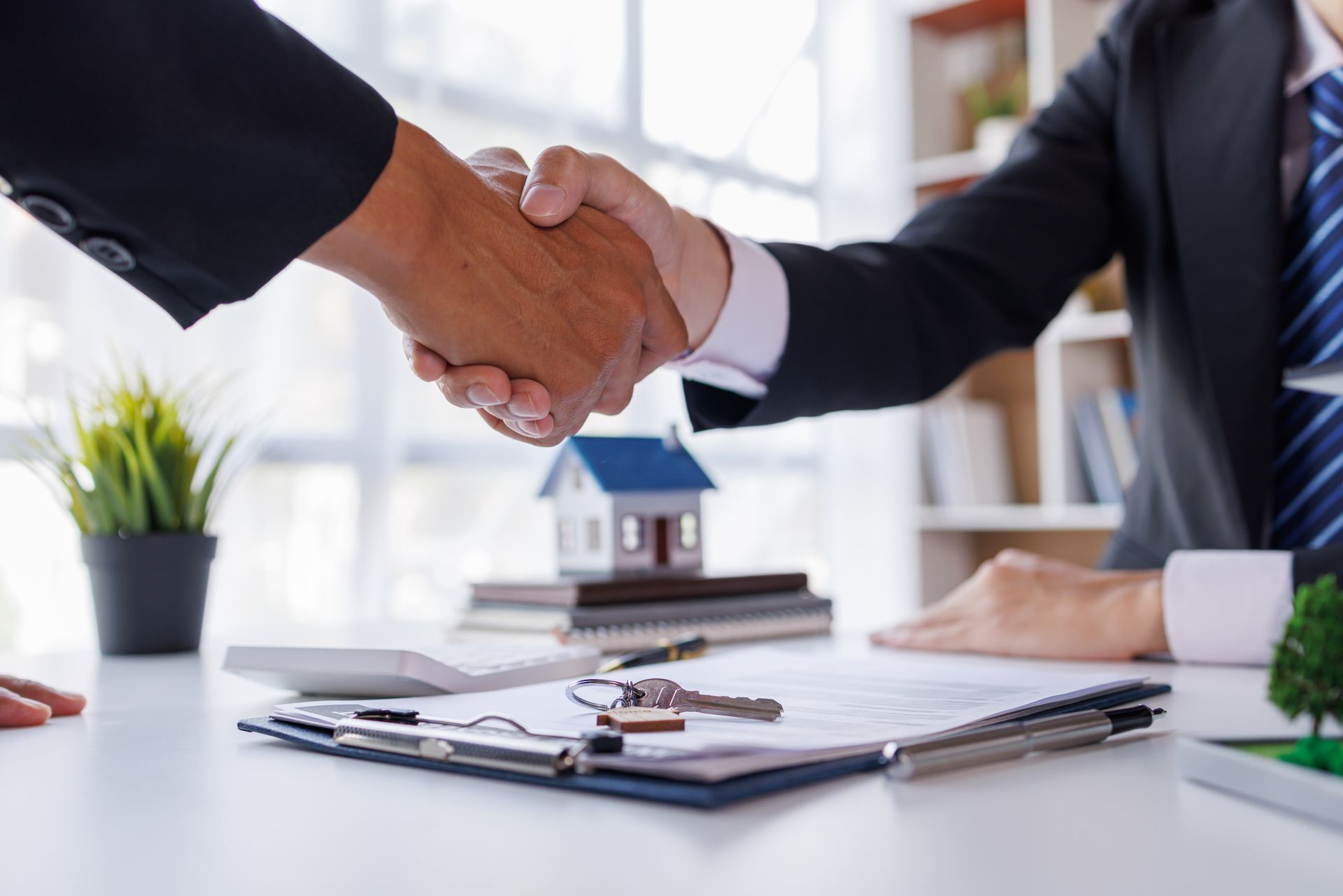 Two professionals in business suits shake hands over a desk with a small house model, documents, and keys. Two professionals in business suits shake hands over a desk with a small house model, documents, and keys.