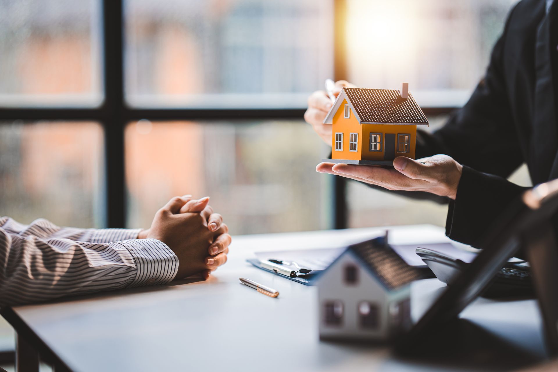A person in a business suit holding a small model house over a desk, with a client seated opposite. A person in a business suit holding a small model house over a desk, with a client seated opposite.