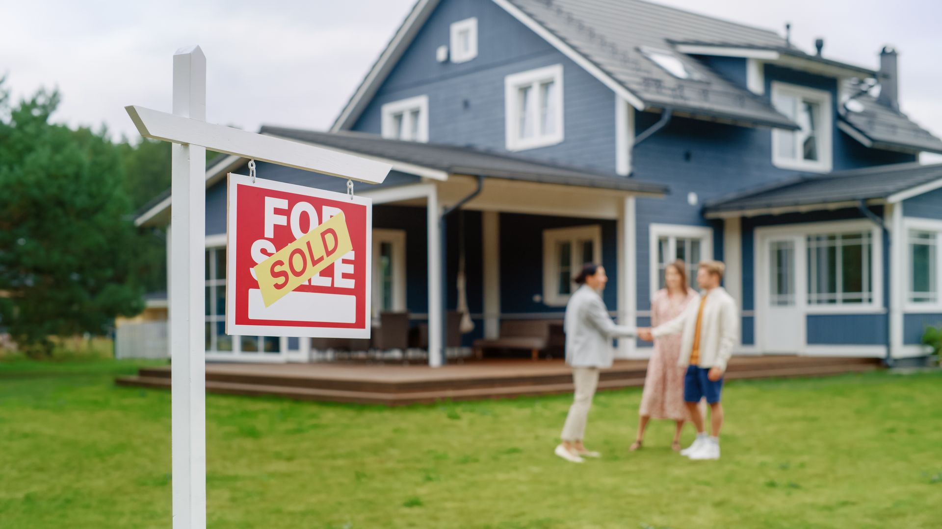 A real estate agent shakes hands with a couple in front of a blue house with a A real estate agent shakes hands with a couple in front of a blue house with a