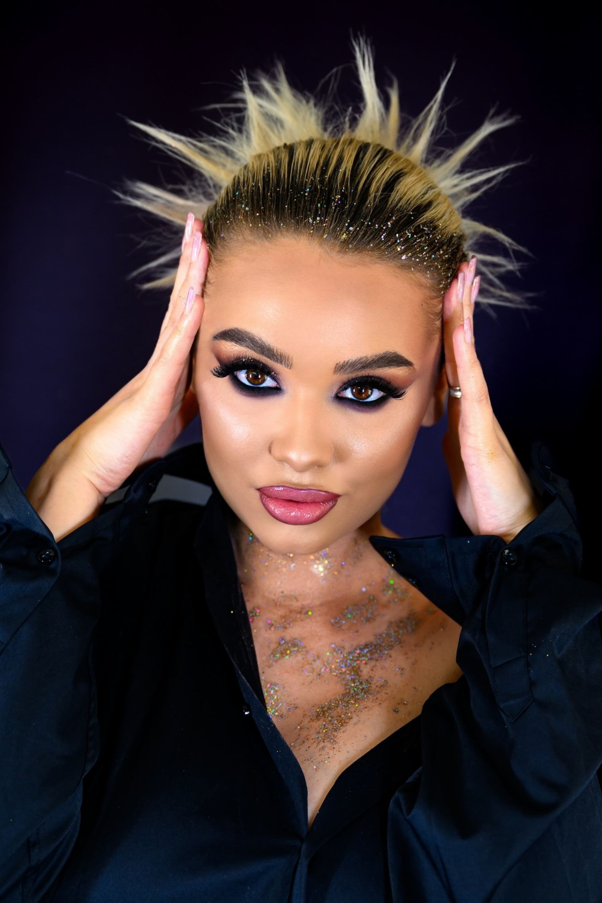 Frau mit stacheligem blondem Haar, dunklem Augen-Make-up und Glitzer, Hände auf dem Kopf, trägt ein schwarzes Hemd.