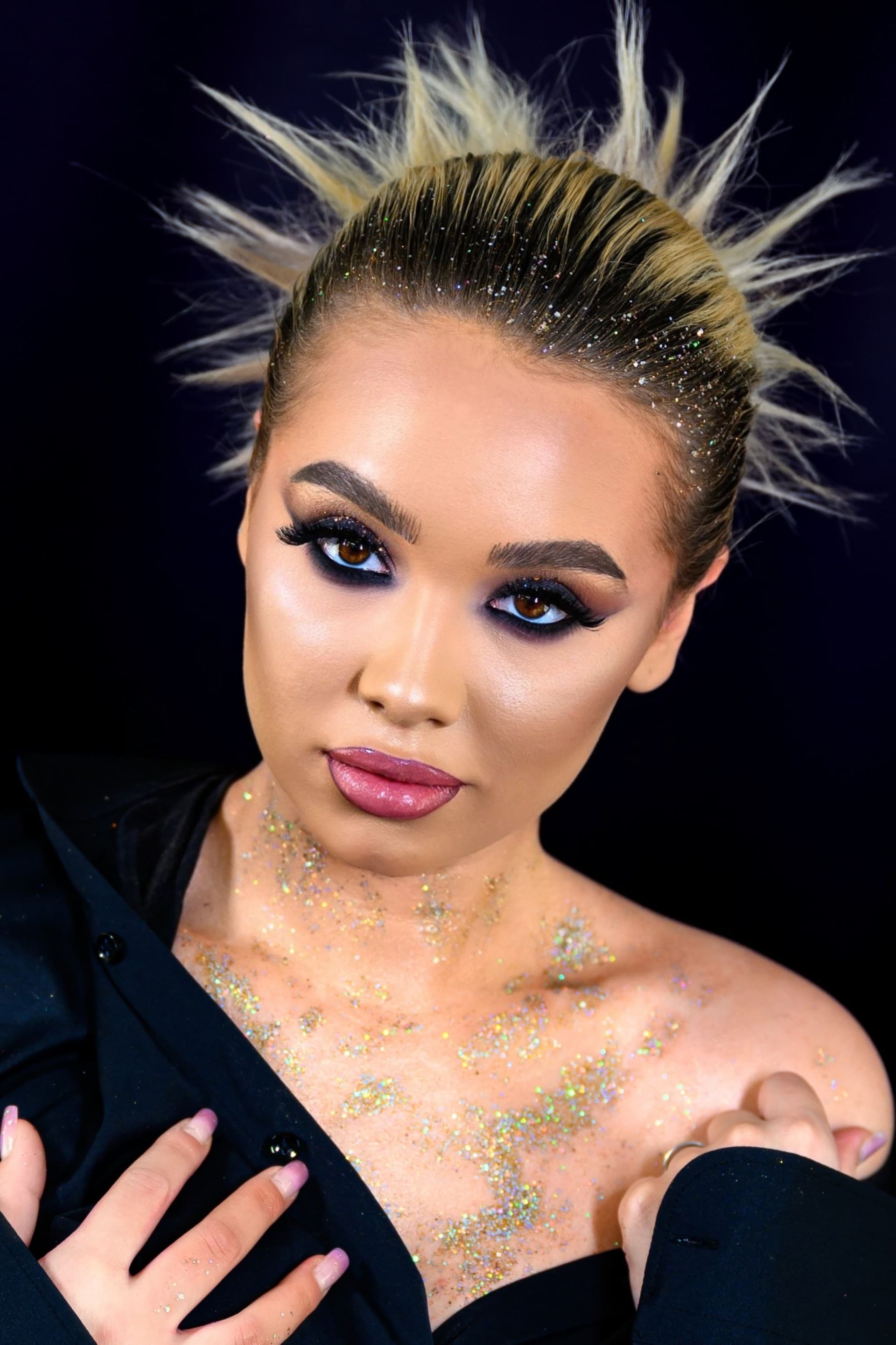 Frau mit stacheligem blondem Haar, dramatischem Augen-Make-up, Glitzer und dunkler Jacke vor schwarzem Hintergrund.