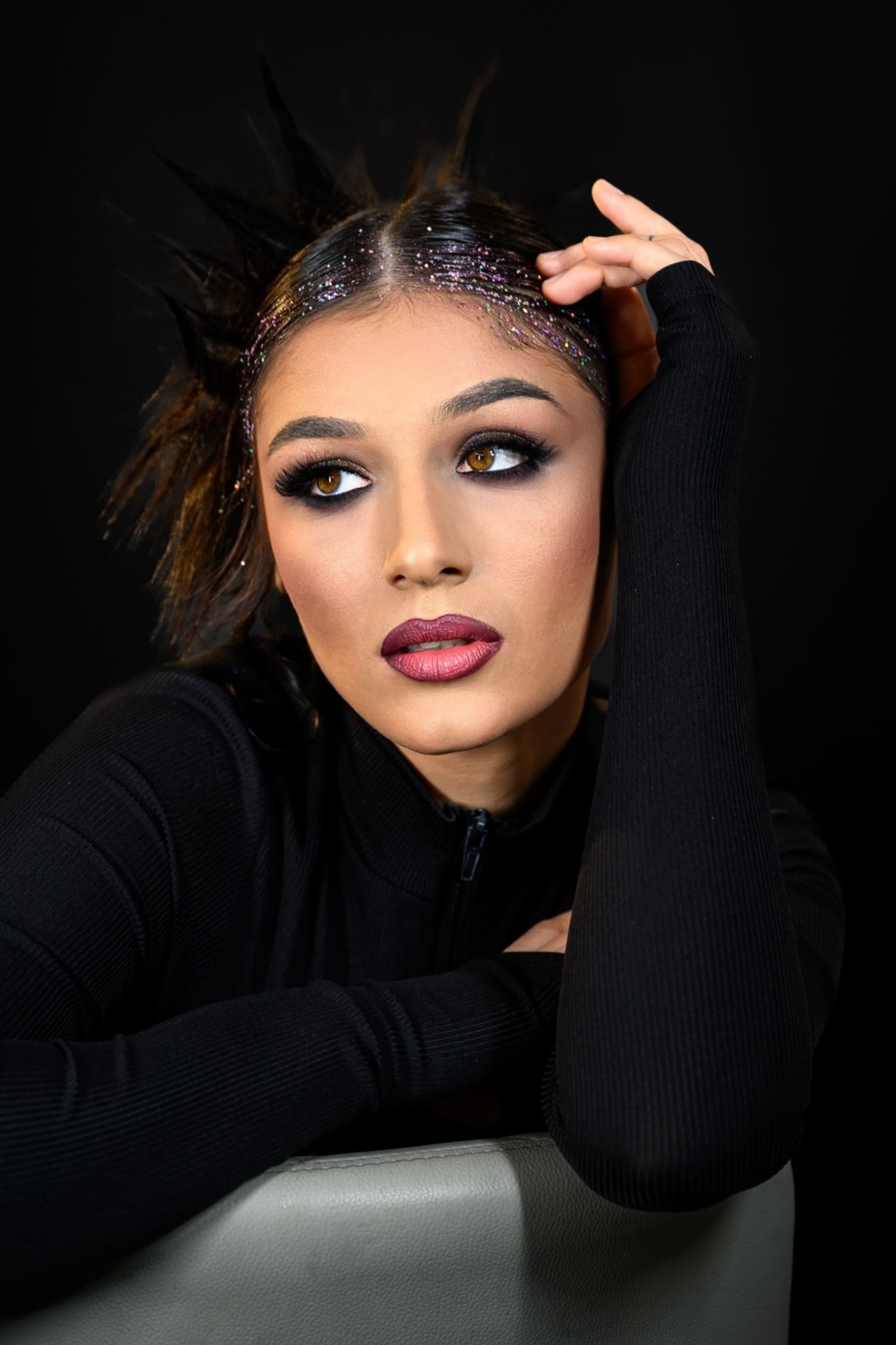 Frau mit dramatischem Augen-Make-up, mit Glitzer gestyltem Haar, schwarzem Oberteil, auf einem Stuhl gelehnt.