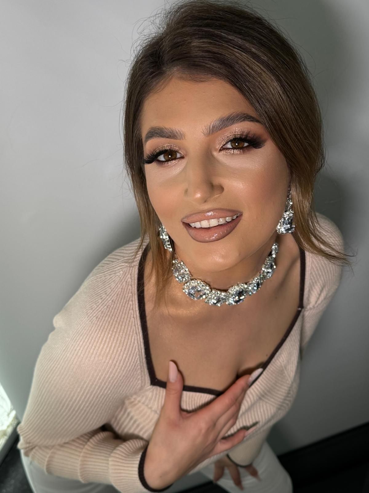 Frau mit glamourösem Make-up und Schmuck lächelt, die Hand auf der Brust, vor einer grauen Wand.