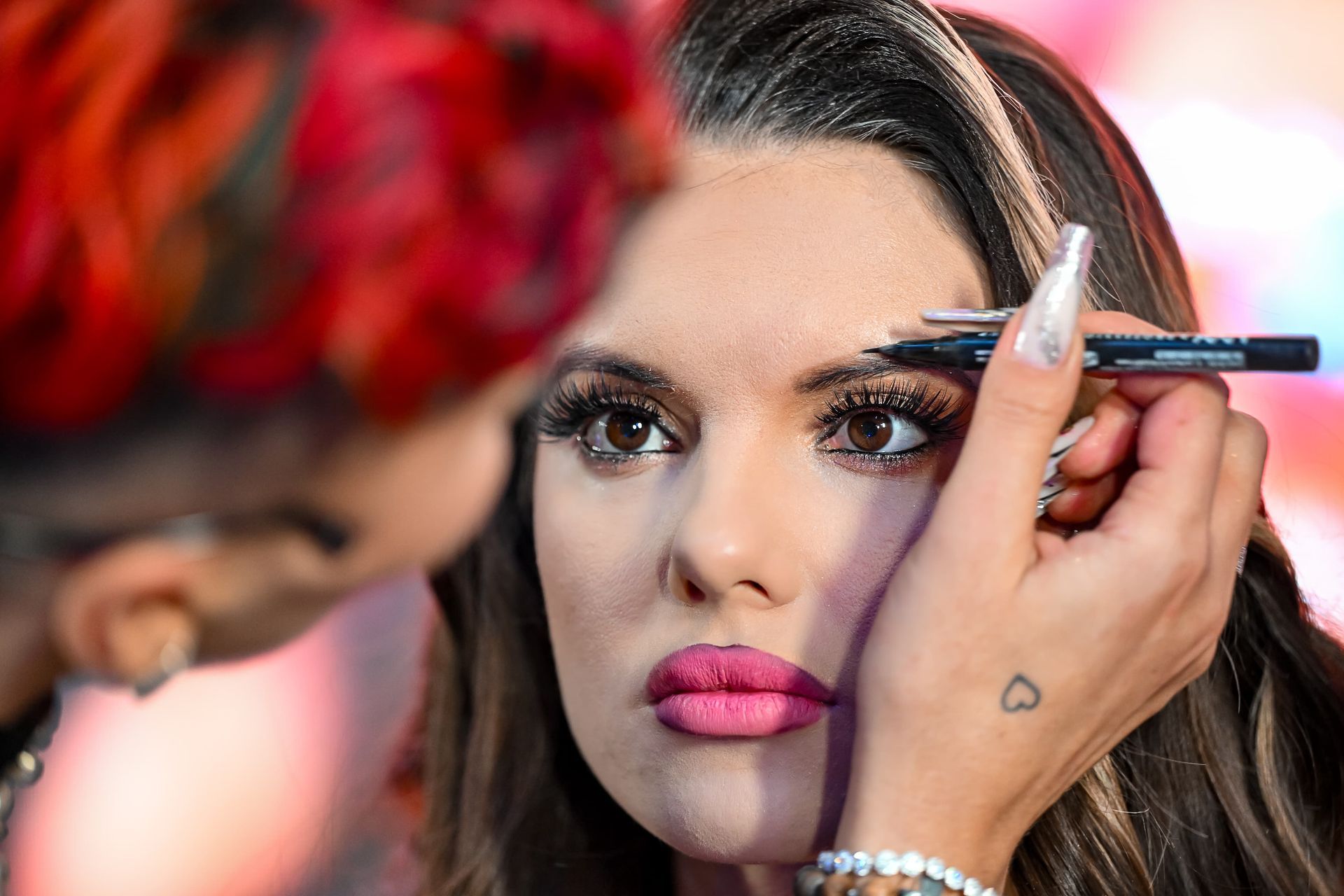 Maskenbildnerin zeichnet auf die Augenbraue einer Frau. Die Frau ist voll geschminkt, hat rosa Lippenstift und lange Nägel.