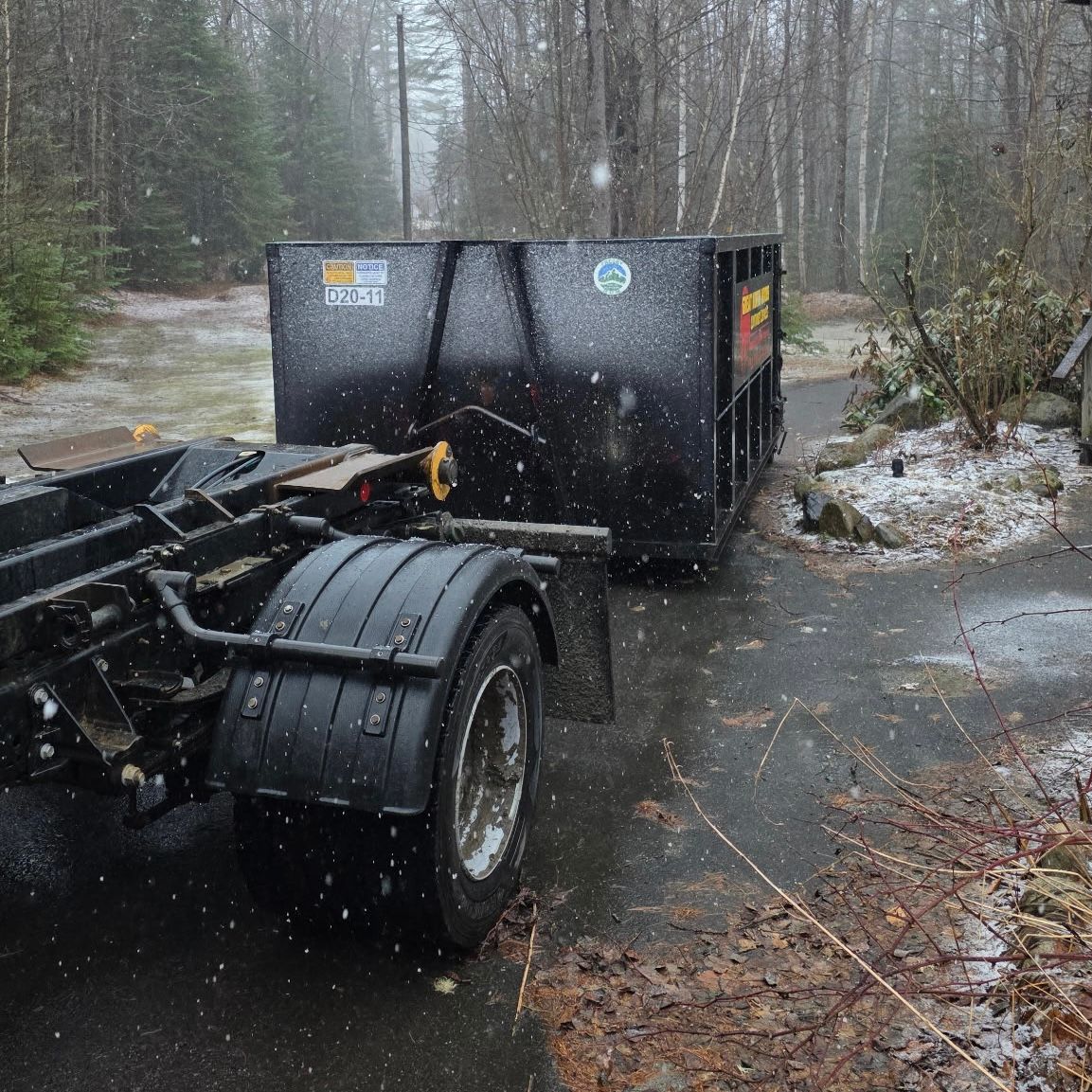 A black dumpster on a truck bed in a snowy driveway. Trees in the background.