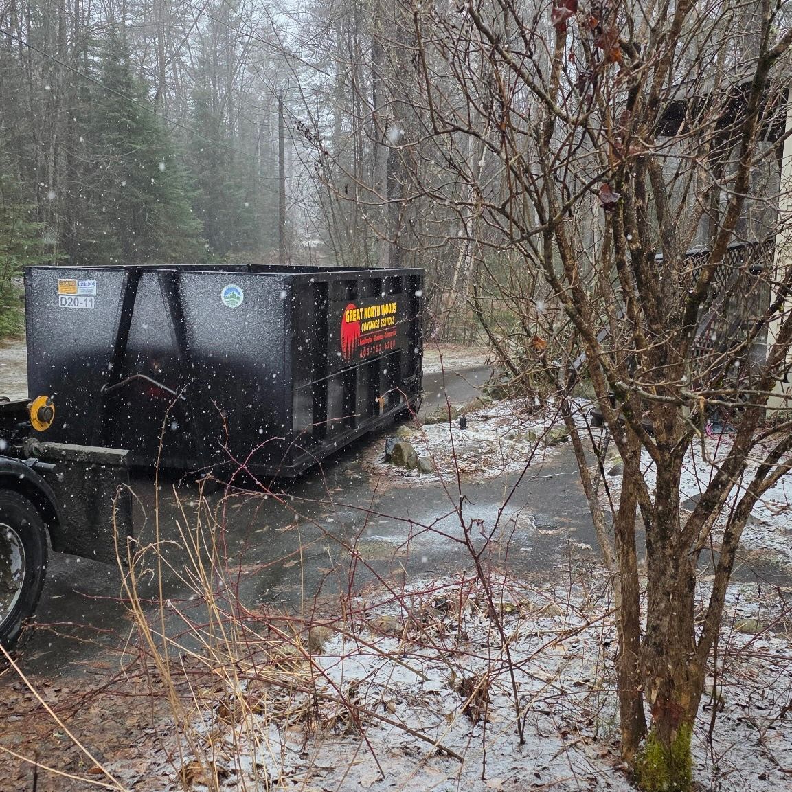 Black dumpster on truck parked on snowy driveway, bare trees in foreground, forest in background.
