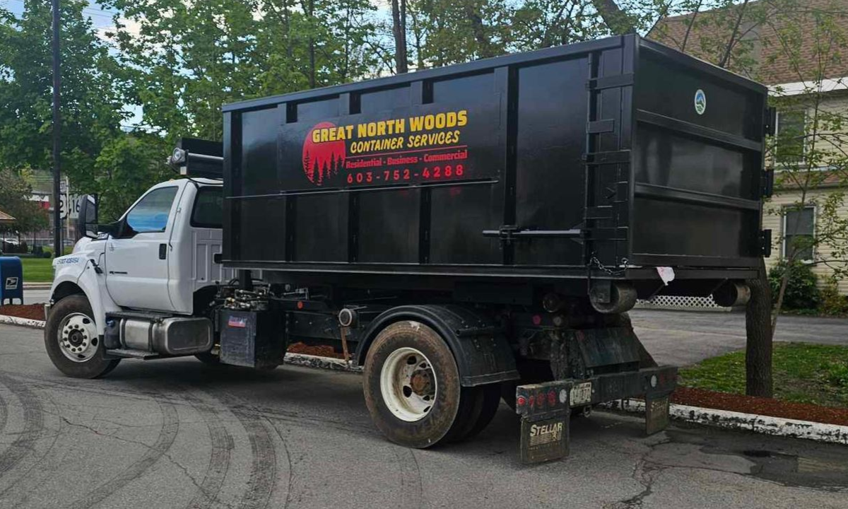 White truck with black dumpster body parked on a street beside trees and apartments