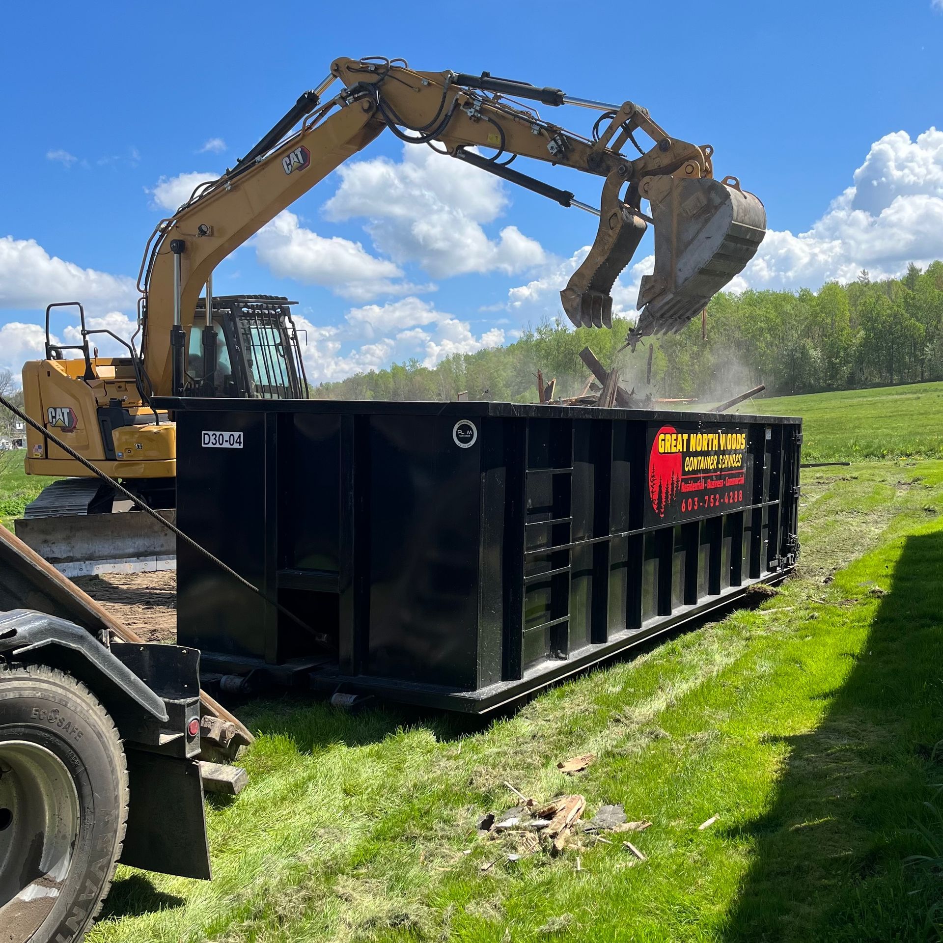 An excavator loading debris into a large black dumpster on green grass.
