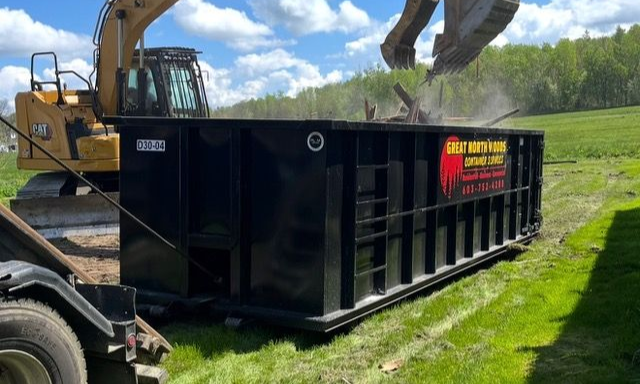 Black construction dumpster beside excavator on grassy field under a blue sky