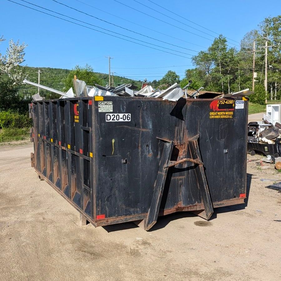 Black dumpster filled with metal scraps on a gravel surface, under a clear blue sky.