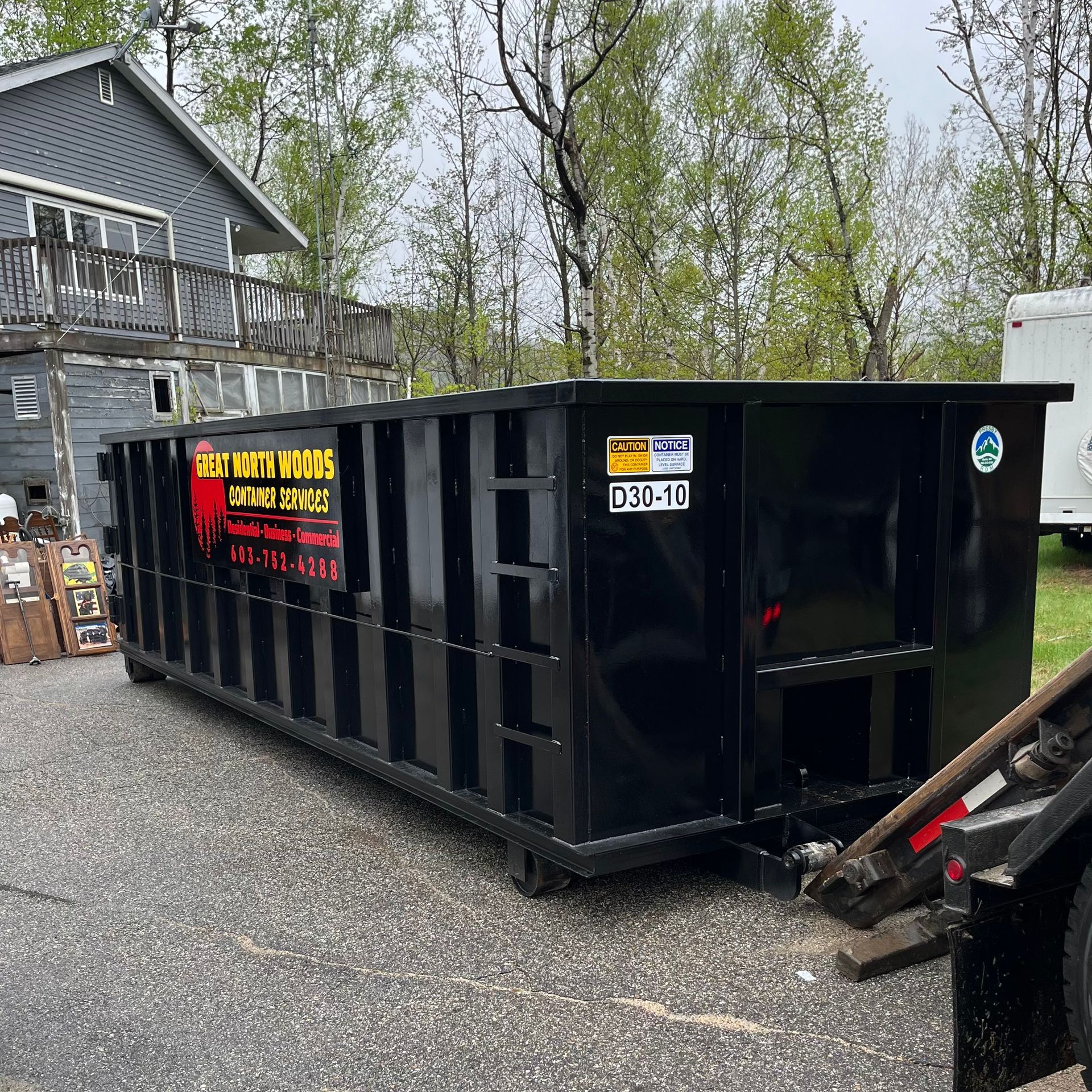 Black dumpster parked on asphalt near a house and a truck.